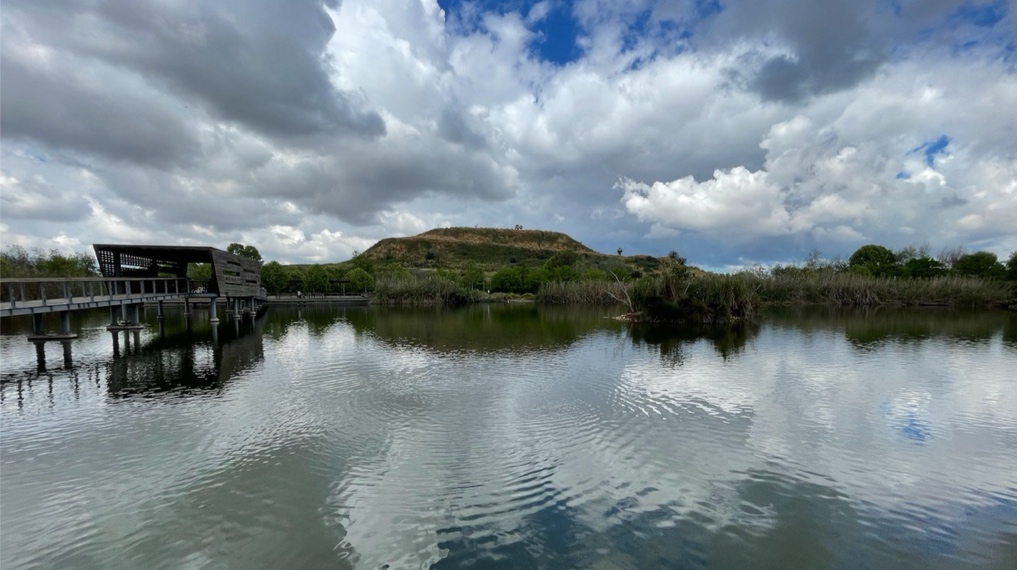 מראה מהשביל המעגלי בחלקו הצפוני - האגם בפארק האקולוגי בהוד השרון, הגשר החוצה אותו, הפרגולה במרכזו והר הזבל בולט מדרום